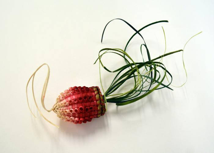 A basket woven in the shape of a radish with a blending of dark pink at the top to a white at the bottom. The cover is green and gives the impression of leaves.